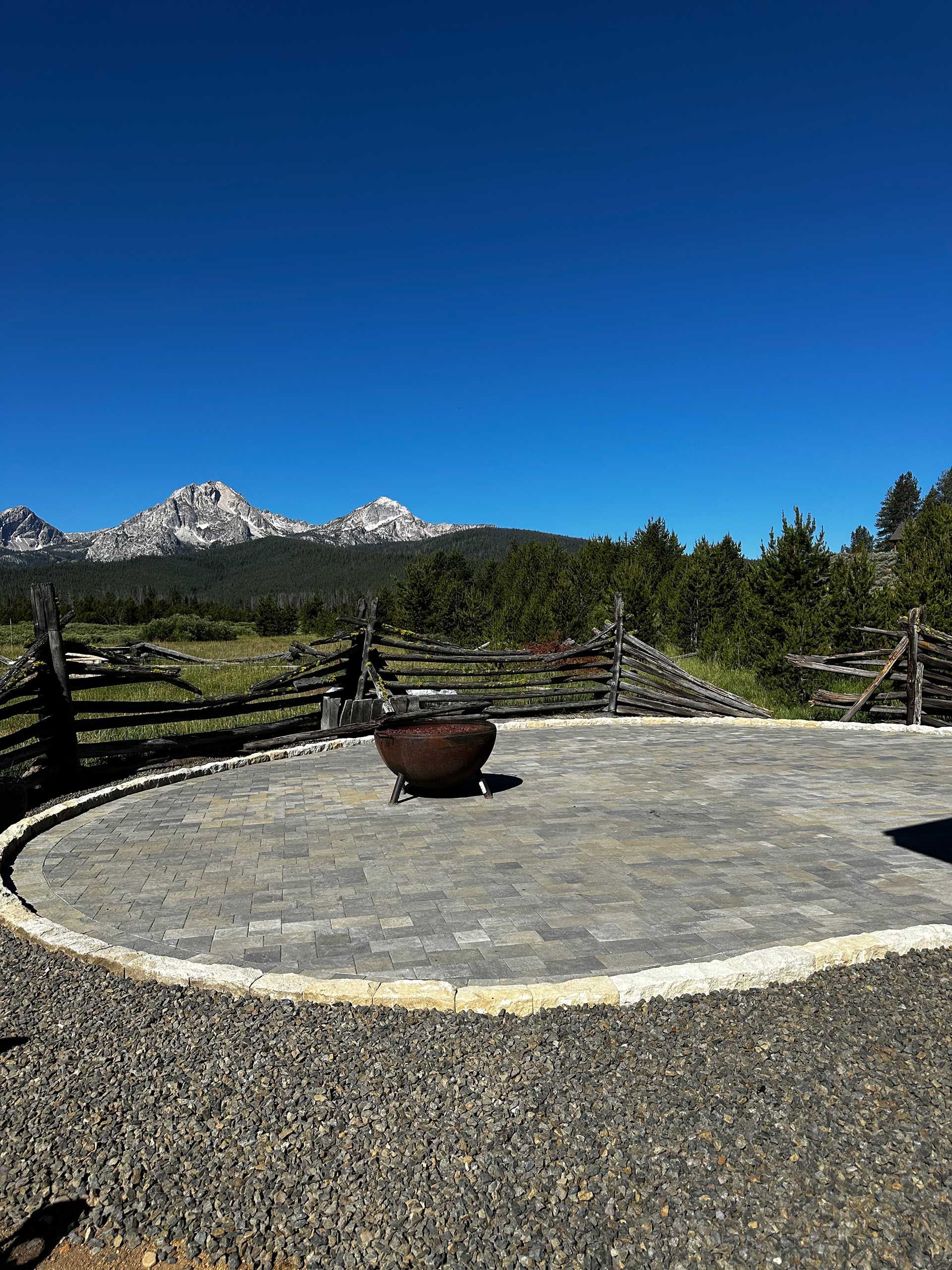 Stone fire pit on a gravel patio with mountain peaks and evergreen trees under a clear blue sky