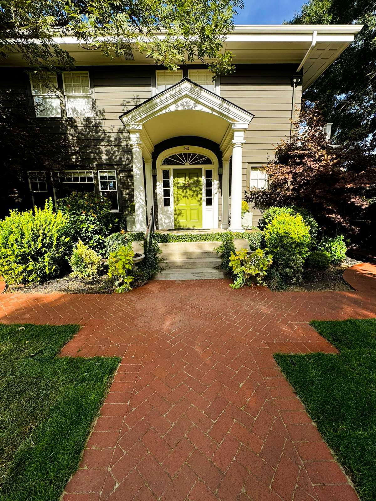 Brick walkway leading to a house entrance with a white arched porch and green front door, framed by shrubs.
