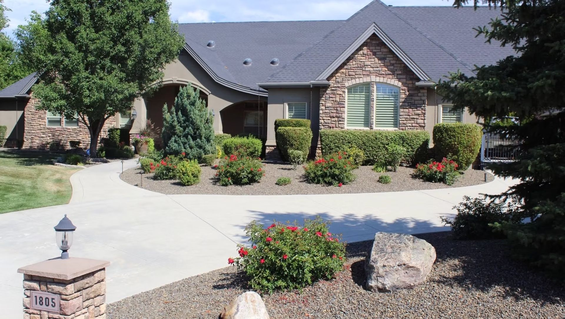 Brick suburban house with landscaped front yard, driveway, and flower beds under a clear sky
