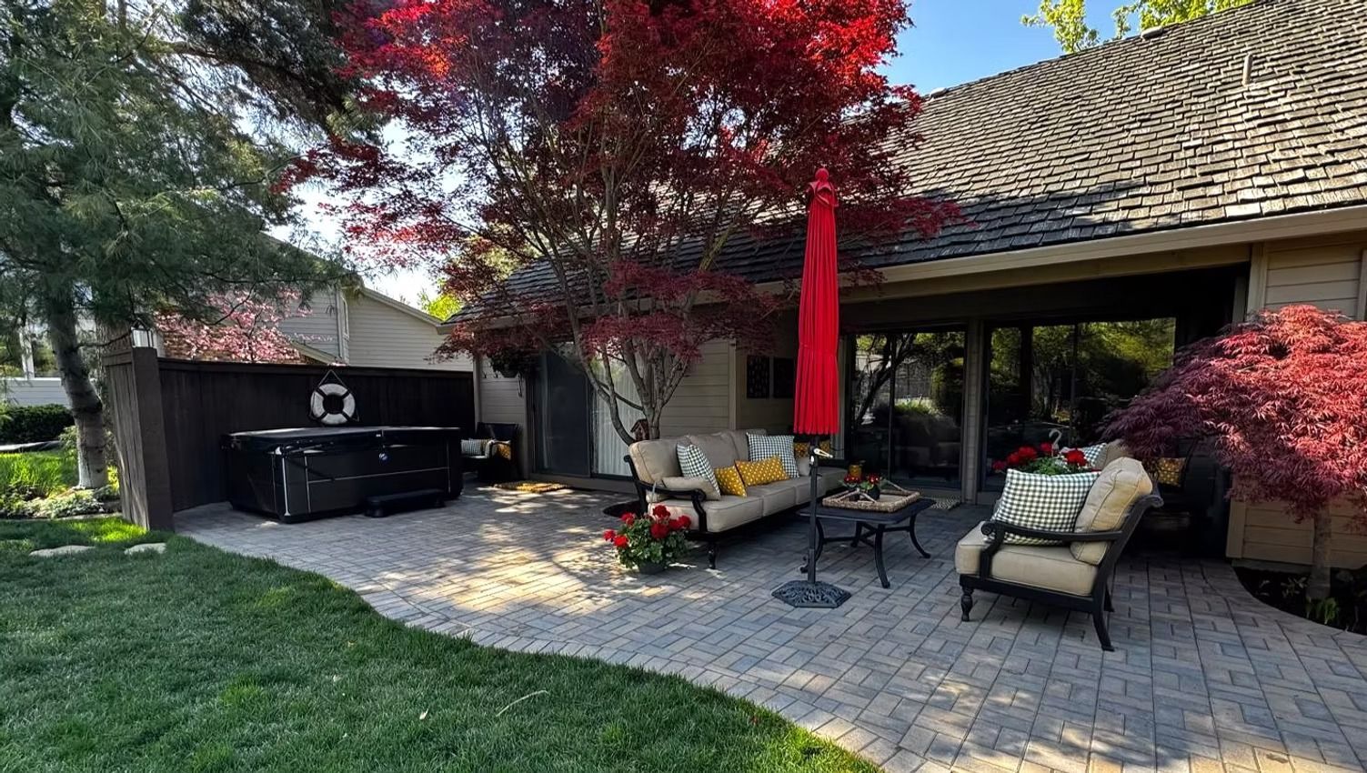 Back patio with lounge chairs, grill, and red umbrellas beside a brick house and maple trees.