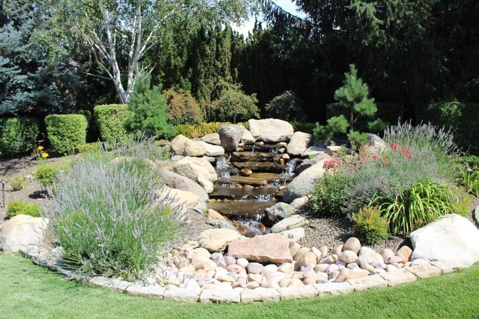 Rock garden with a small cascading water feature, surrounded by flowers, stones, and green shrubs.