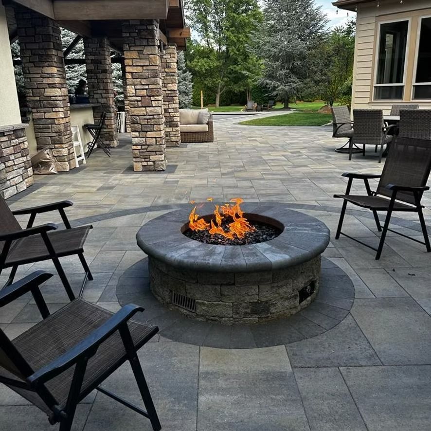 Stone patio with a circular fire pit burning, surrounded by black chairs near a stone house.
