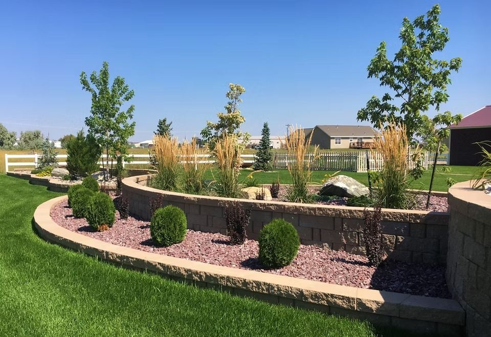 Landscaped suburban garden with stone retaining wall, shrubs, young trees, and green lawn under a blue sky
