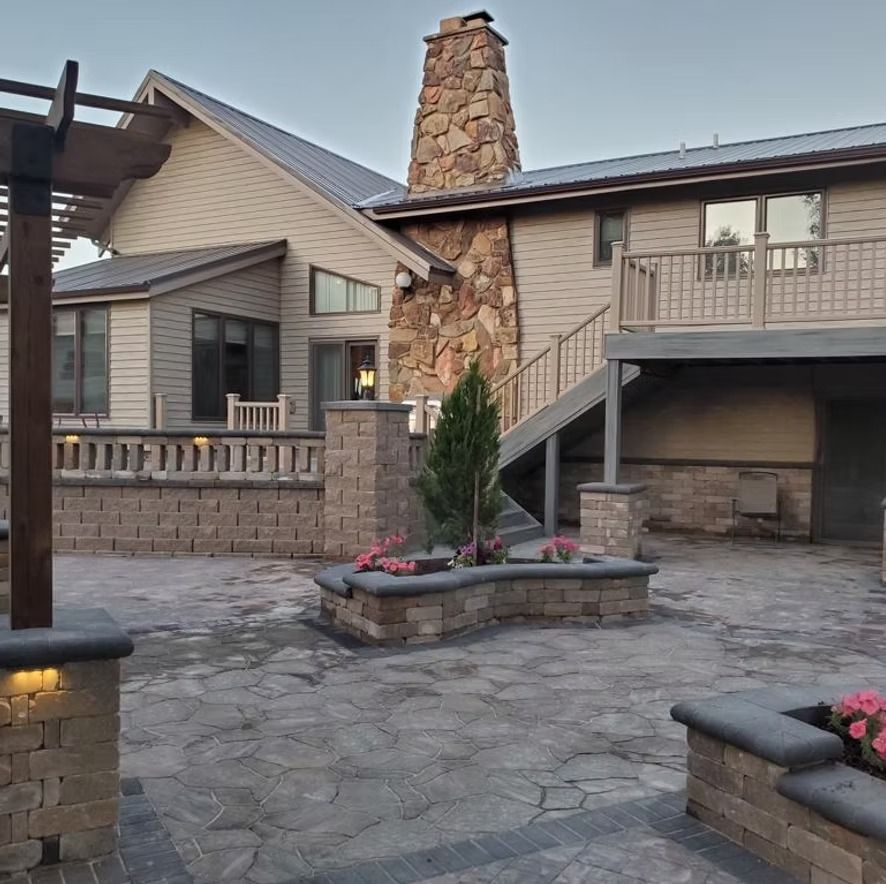 Stone courtyard with low planters, stairs, and a chimney beside a beige house at dusk