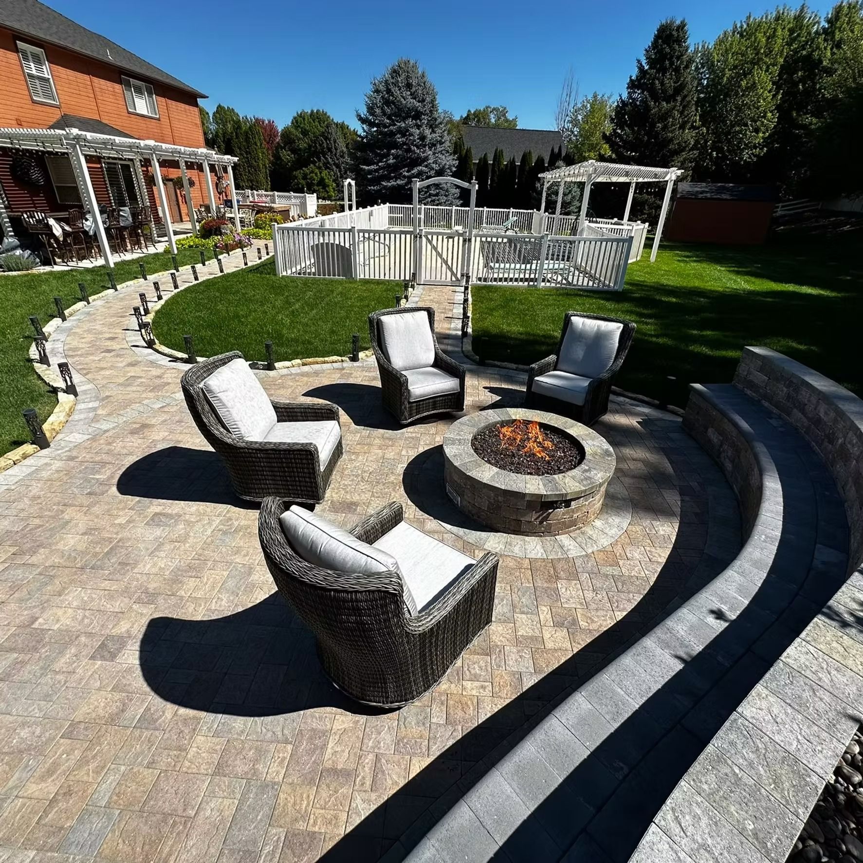 Backyard patio with circular fire pit, four wicker chairs, and curved stone seating wall under a clear blue sky
