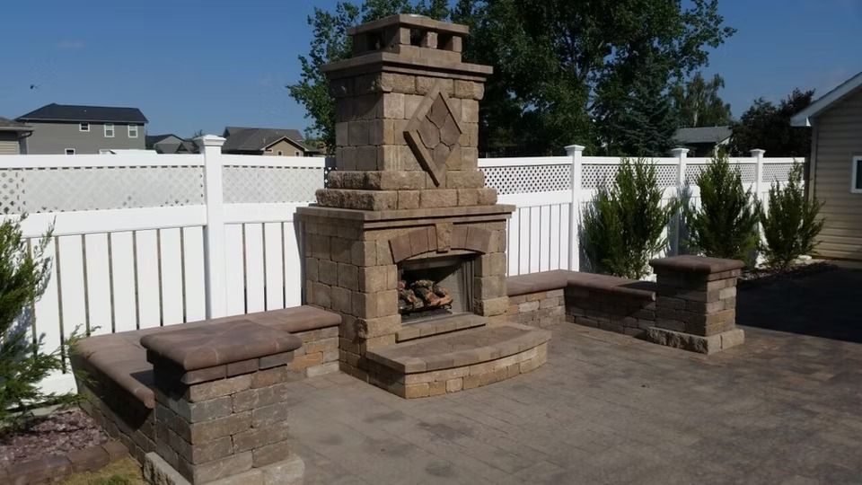 Outdoor stone fireplace with matching benches on a paved patio beside a white fence