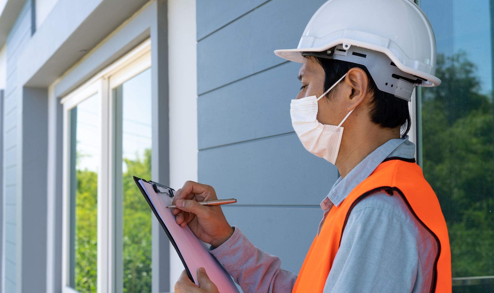 A construction inspector in a hard hat and orange vest wearing a mask, writing on a clipboard outdoors.