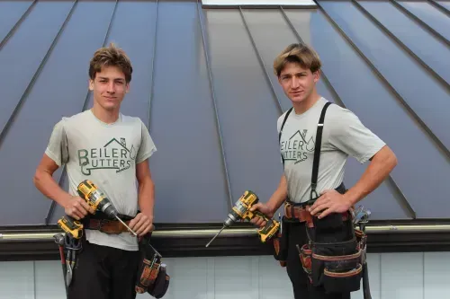 Two men wearing tool belts stand on a roof, holding drills. The sign on their shirts reads "Beiler's Gutters".