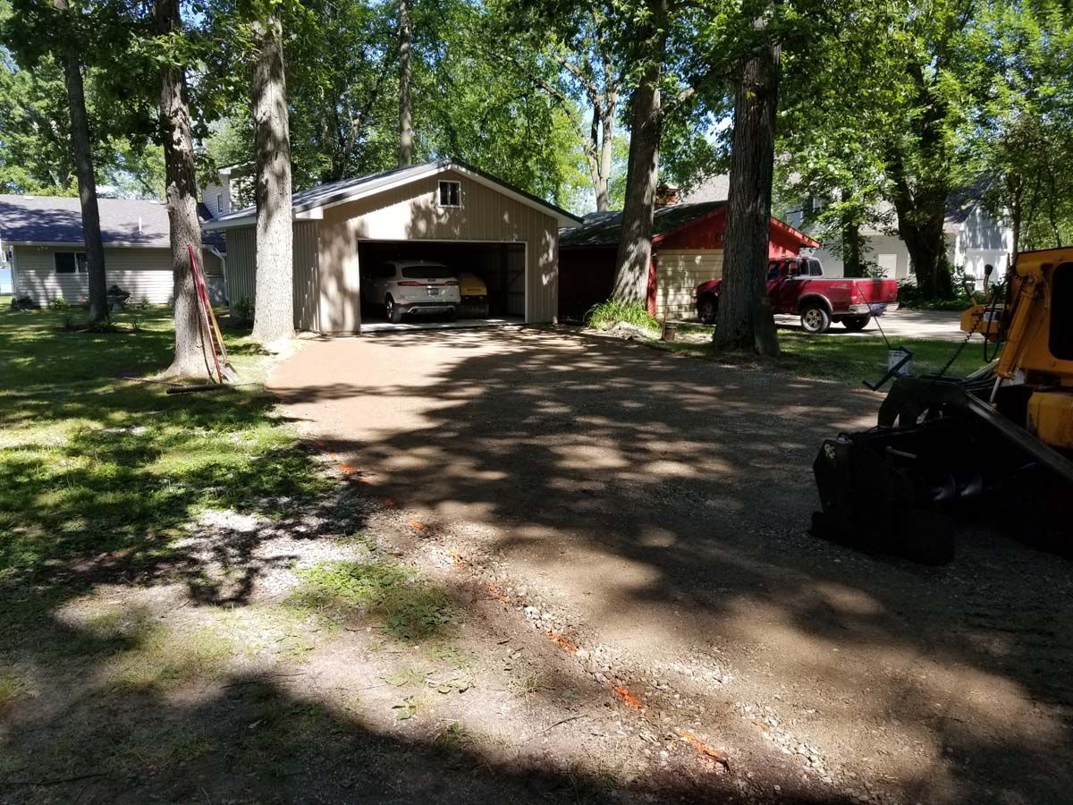Gravel driveway leading to a tan garage with a car inside. Red pickup truck in the background, trees surround.
