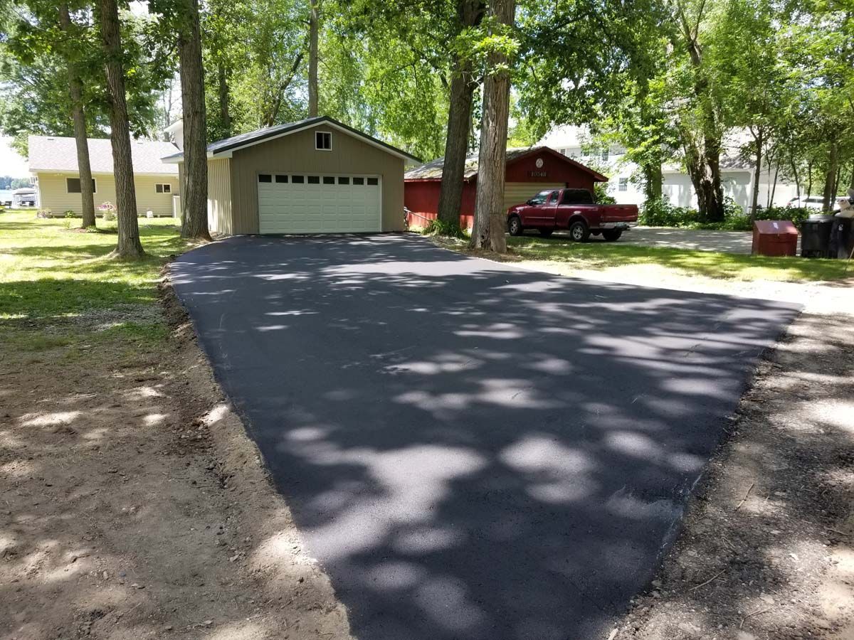 Newly paved asphalt driveway leading to a garage, with trees and a red truck nearby.