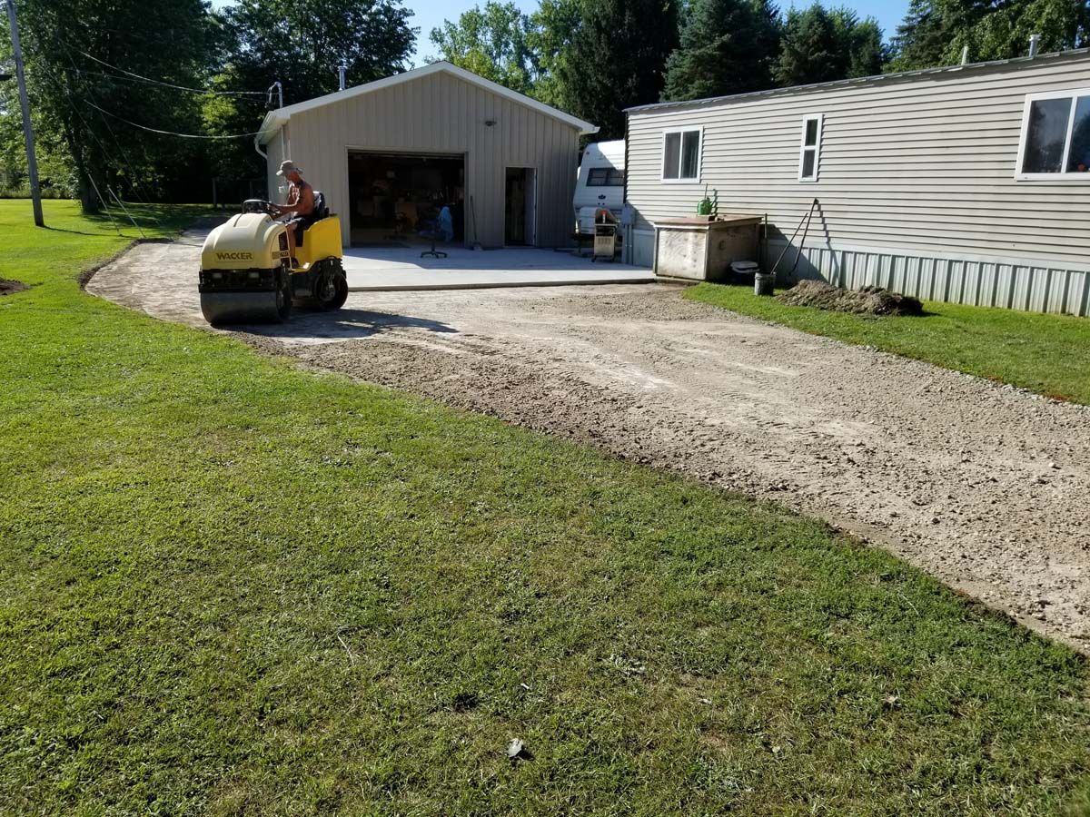 A small roller compacts gravel driveway near a garage and mobile home on a sunny day.