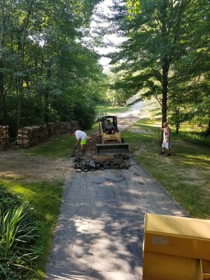 A small tractor and two men work on a driveway surrounded by trees. One man shovels, the other stands.