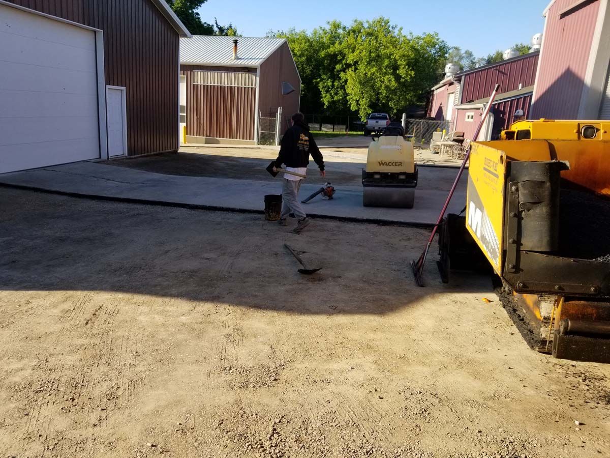 Person leveling asphalt with tools; roller compacting newly paved area near buildings.