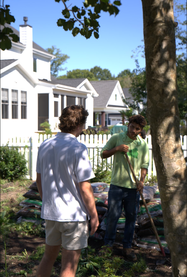 Two people talking in a sunny suburban garden beside white houses and a tree