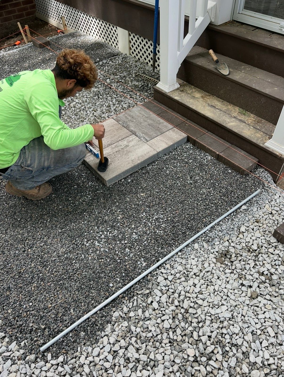Worker kneeling on gravel, laying a stone paver beside outdoor stairs and a white railing.