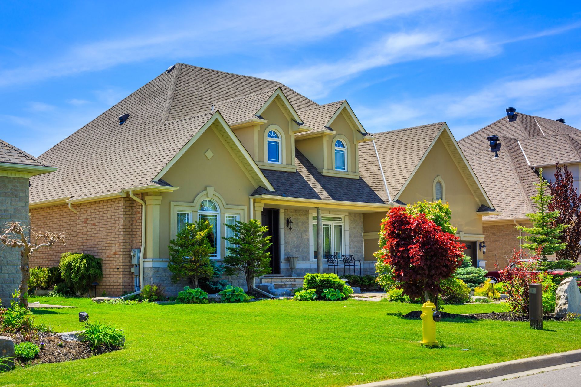 Large brick house with manicured lawn, stone steps, and neatly trimmed hedges under a bright sky