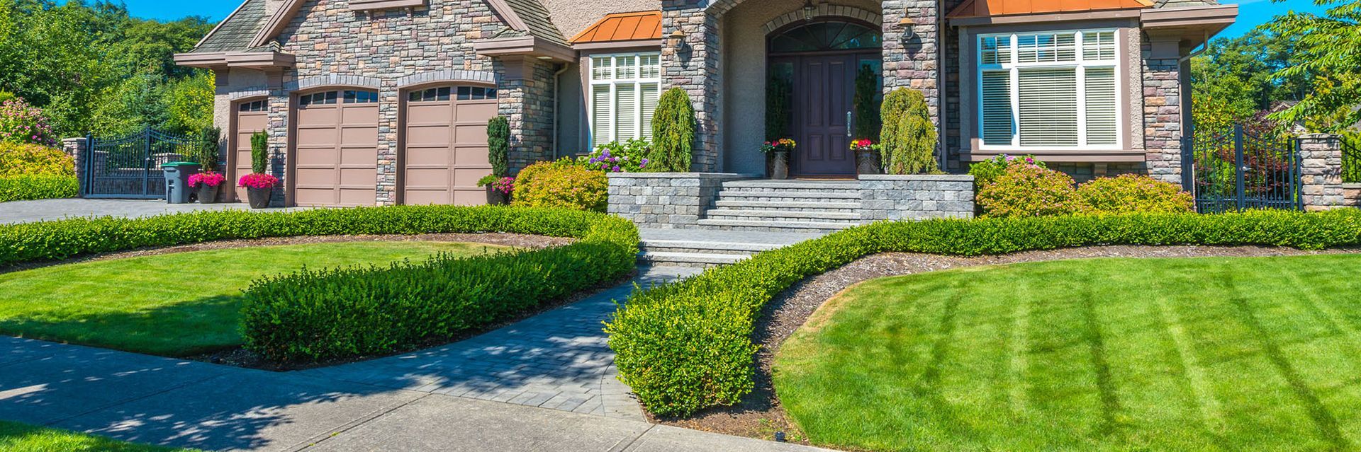 Large brick house with manicured lawn, stone steps, and neatly trimmed hedges under a bright sky