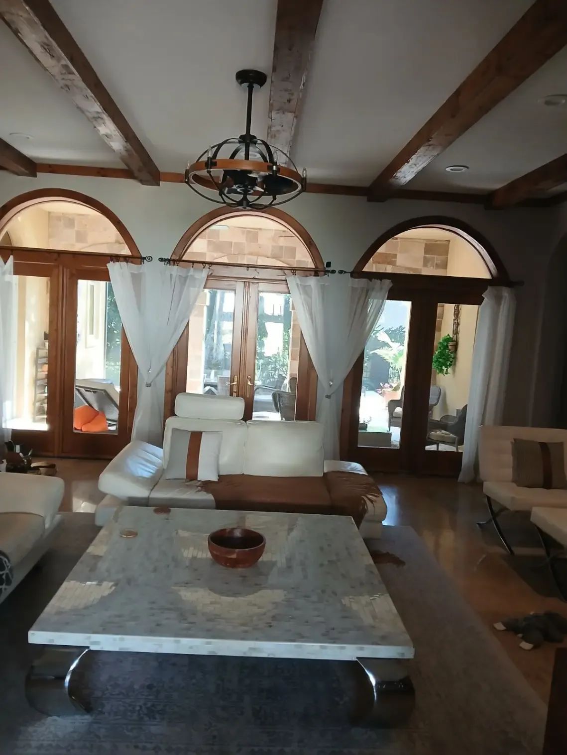 Rustic living room with arched windows, white sofas, and a marble coffee table under exposed wooden beams