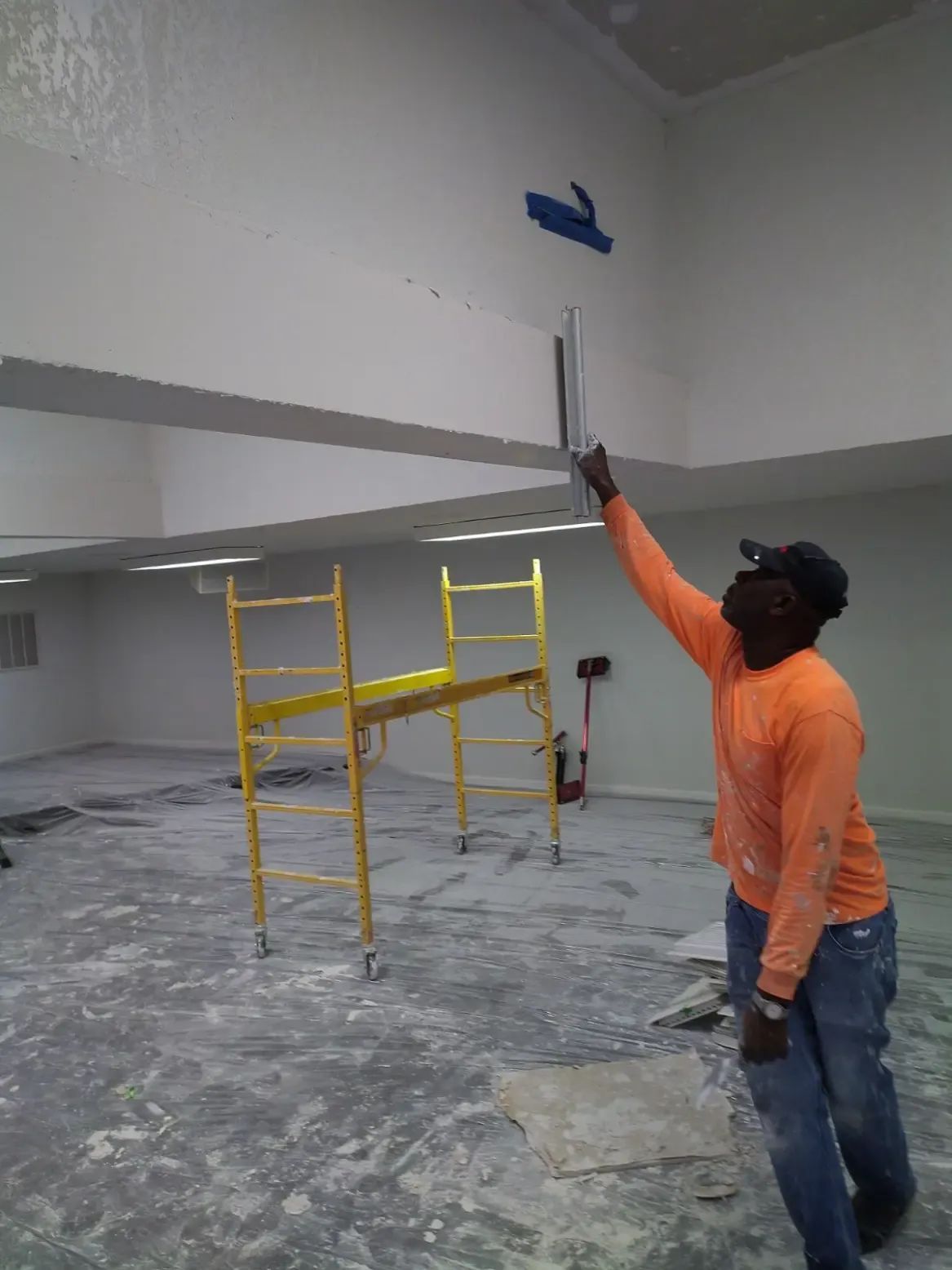 Worker in orange shirt painting a ceiling indoors beside yellow scaffolding.