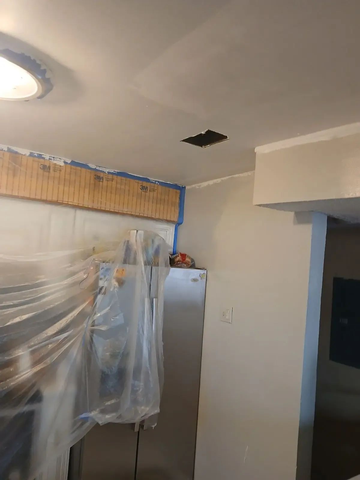 Partially covered kitchen with plastic sheeting, a refrigerator, and a damaged ceiling opening