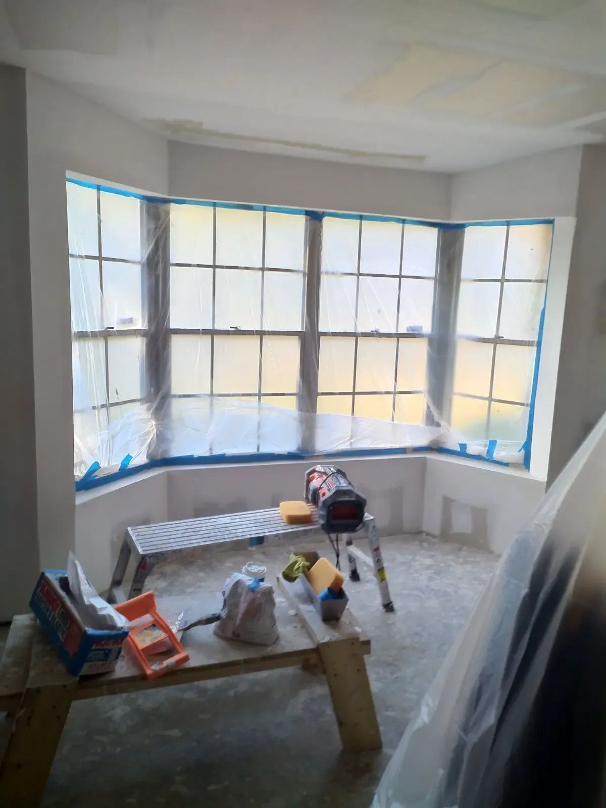 Bright bay window room with painting supplies, drop cloths, and a workbench during renovation