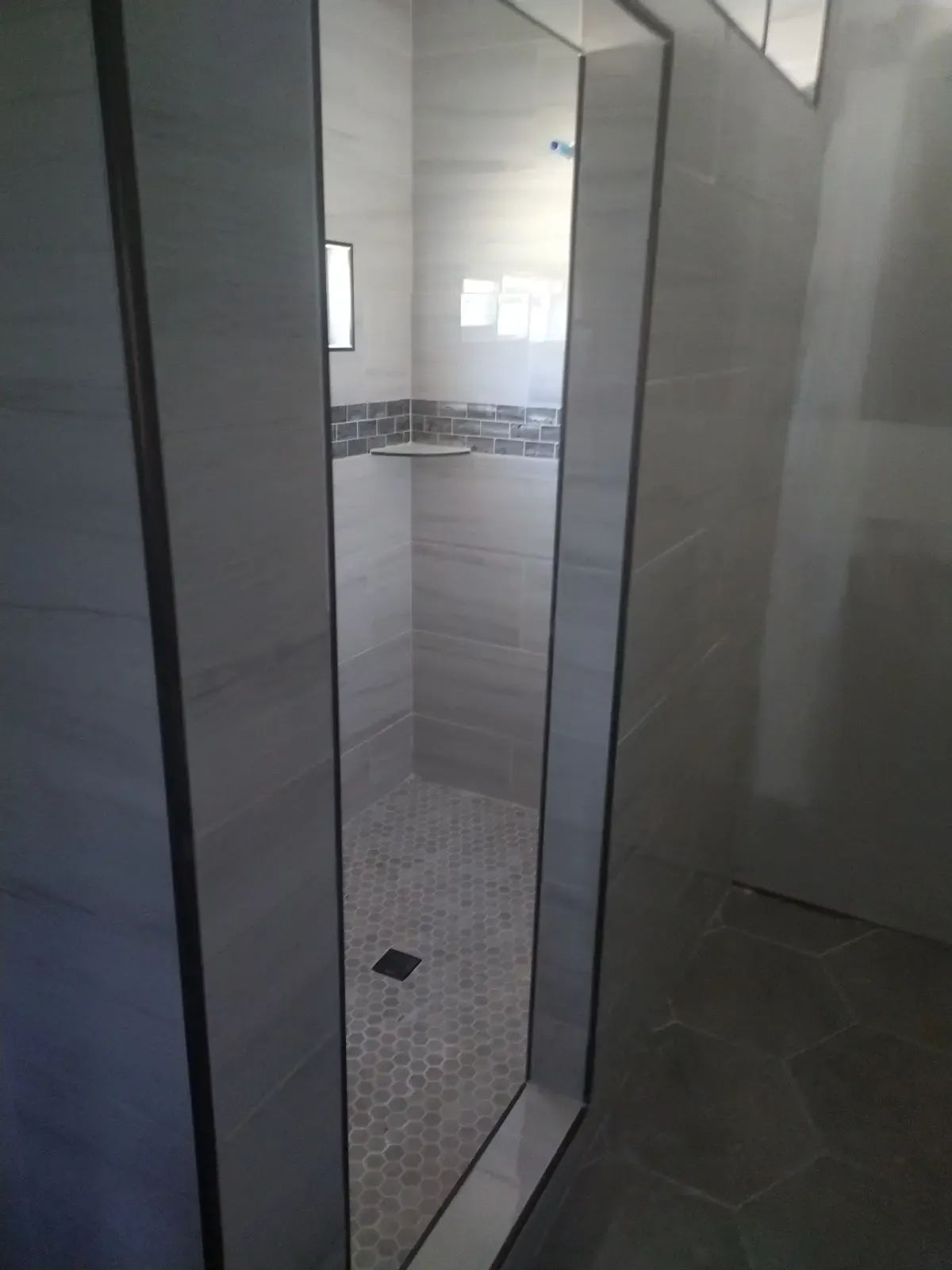 Shower stall with glass door, gray tiled walls, and small mosaic floor tiles