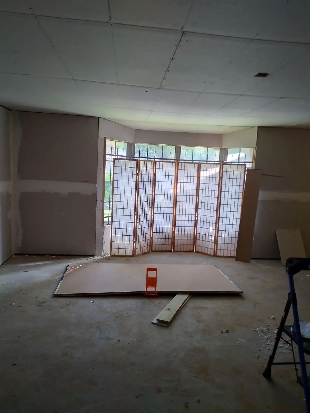 Empty unfinished room with drywall, a folding screen by the window, and construction materials on the floor.