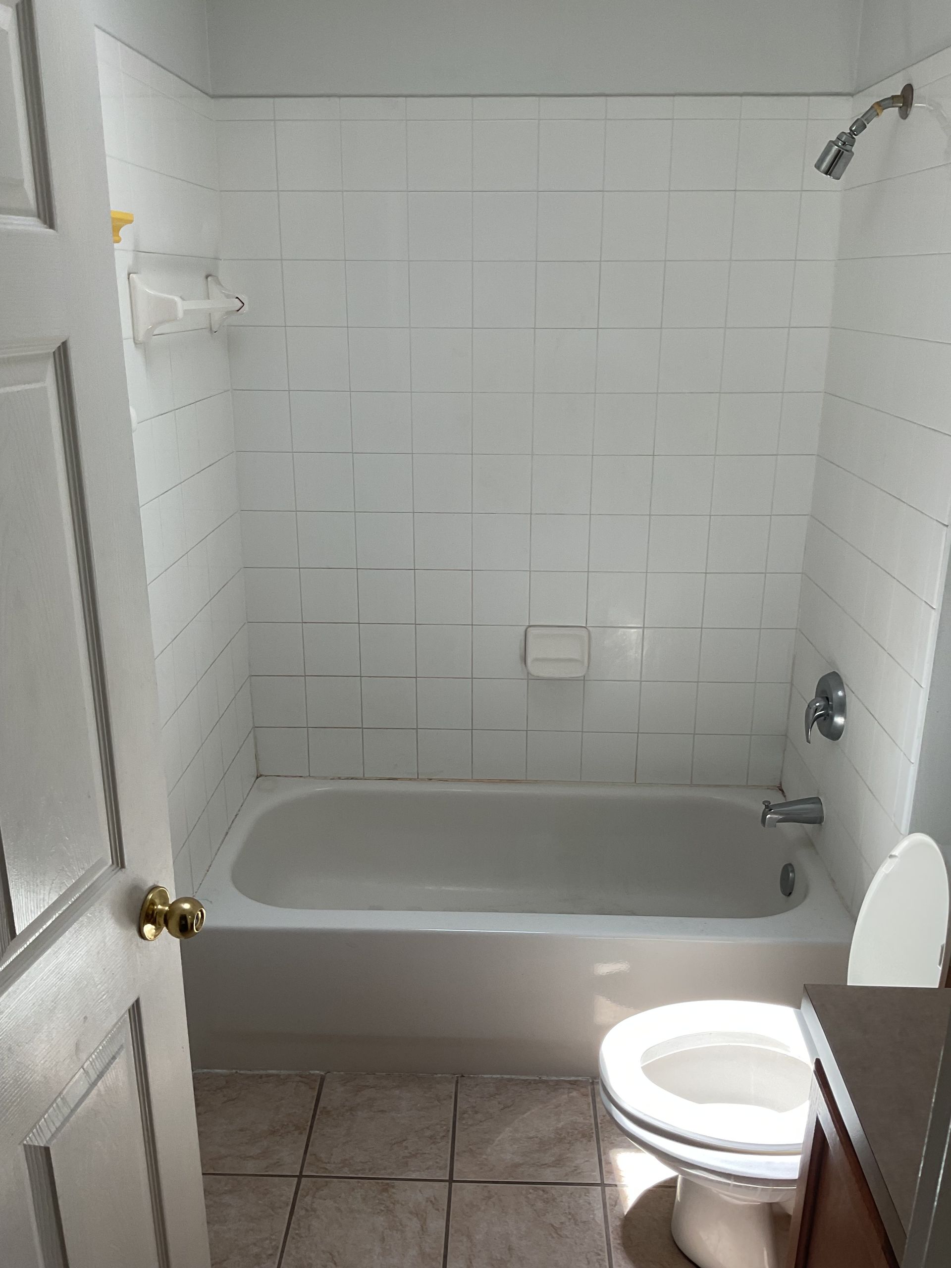 Small bathroom with white tiled shower-tub, beige floor, and toilet in the foreground.