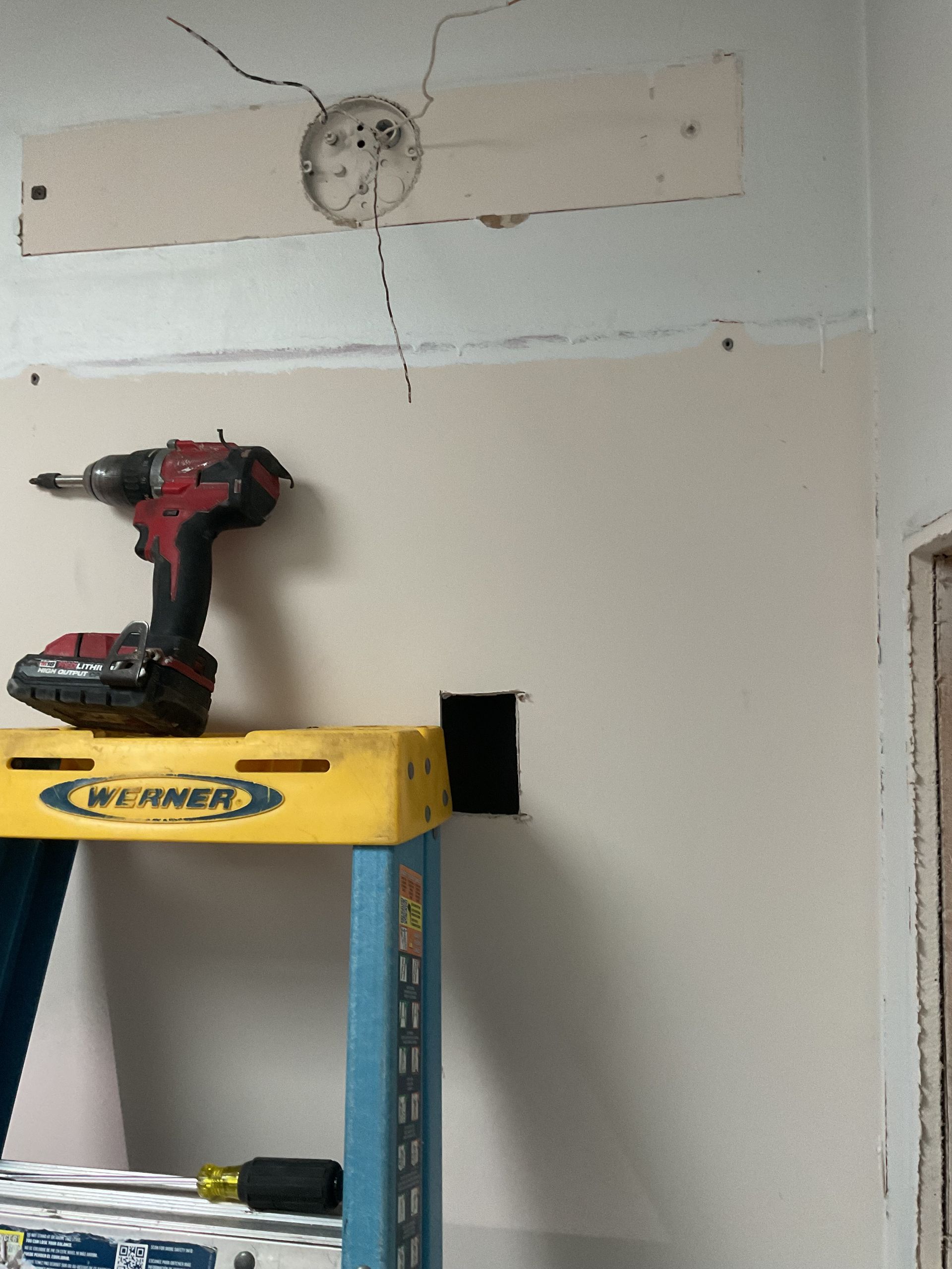 Red cordless drill on a yellow ladder under a wall cutout with exposed wiring and drywall repair patch