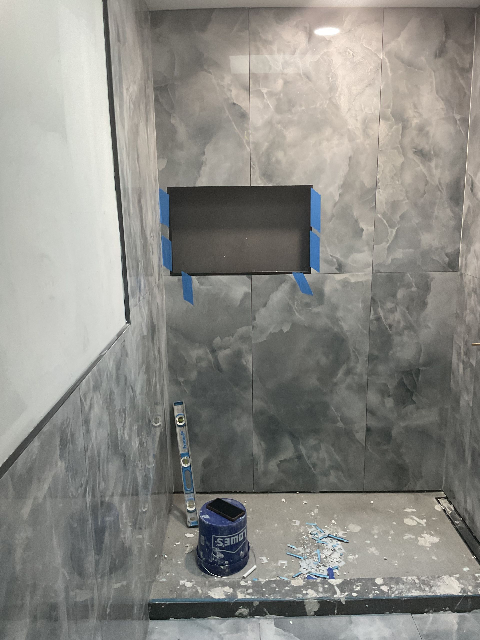 Gray tiled bathroom under renovation with a wall cutout taped in blue and paint supplies on the floor