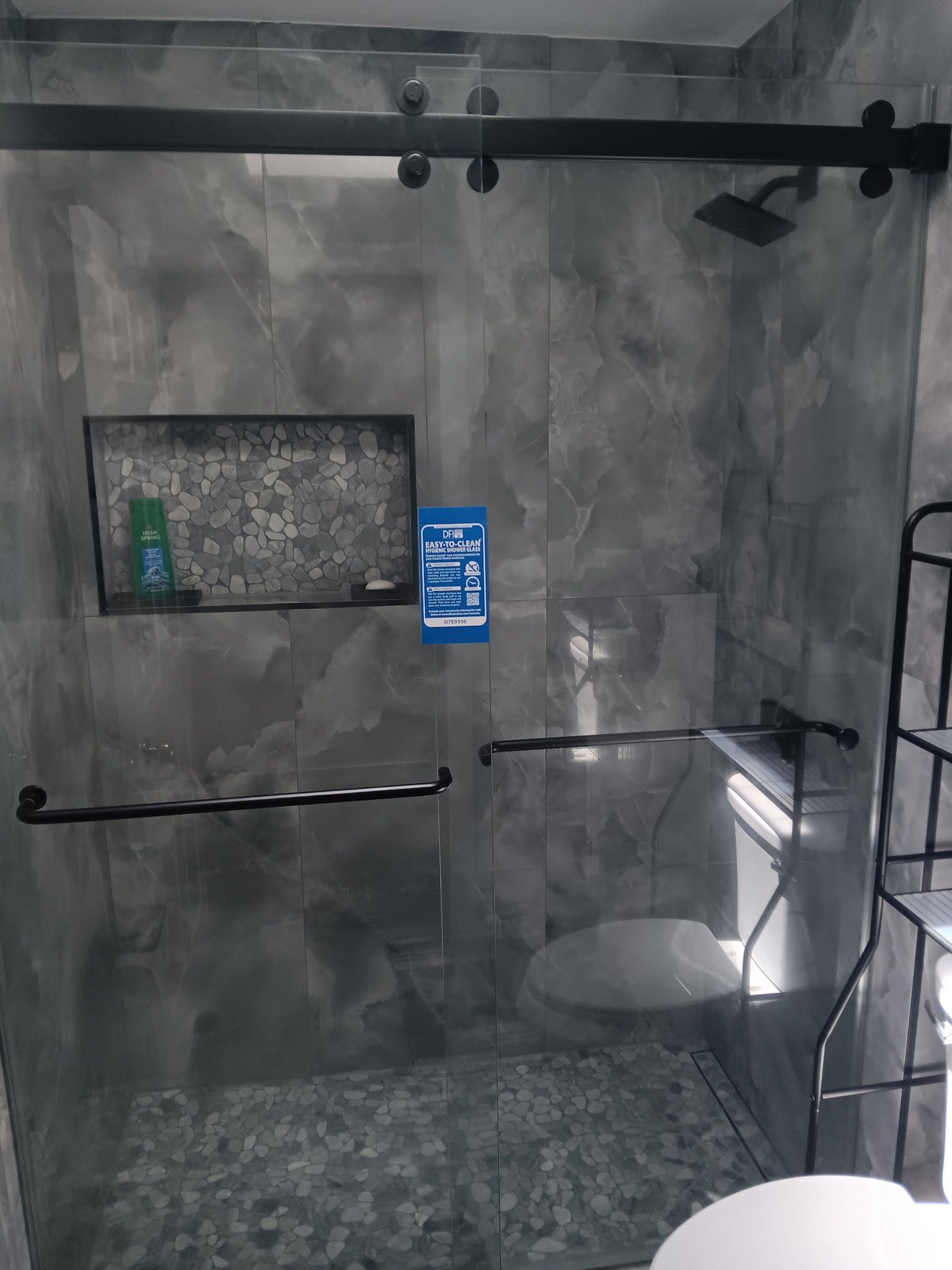Gray tiled shower stall with grab bars, built-in shelf, and a shower chair under a bright light