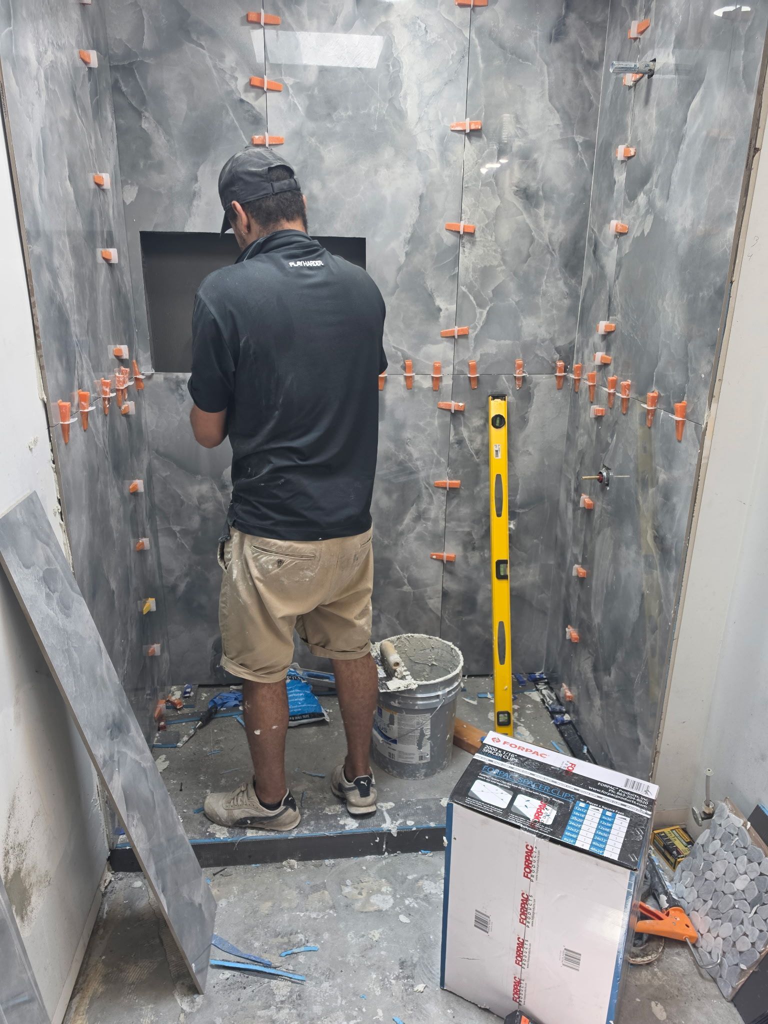 Worker installing wall tiles in a small tiled bathroom, with tools and leveling equipment nearby.
