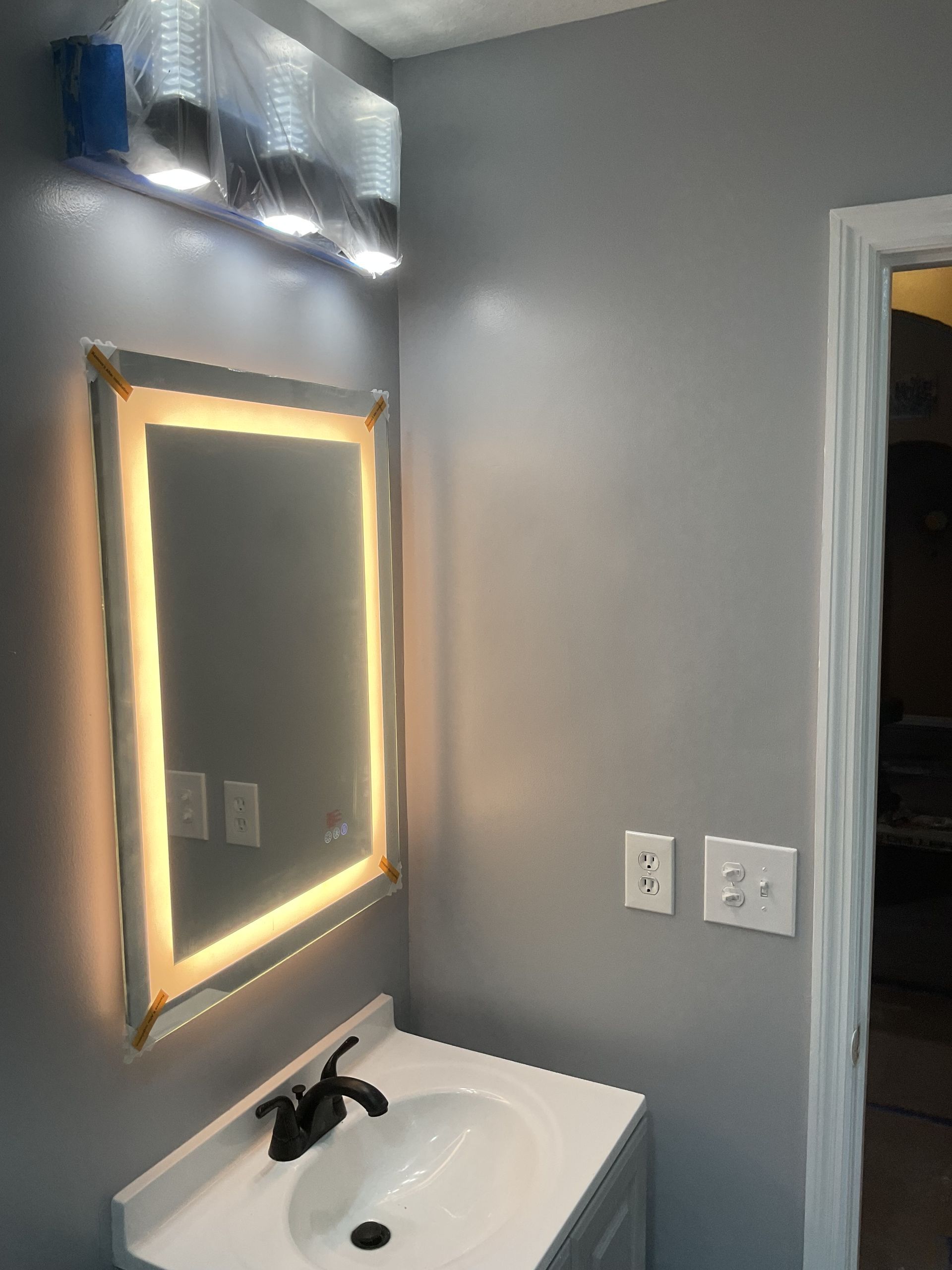 Bathroom vanity with lit mirror, white sink, gray walls, and open doorway to a dark room