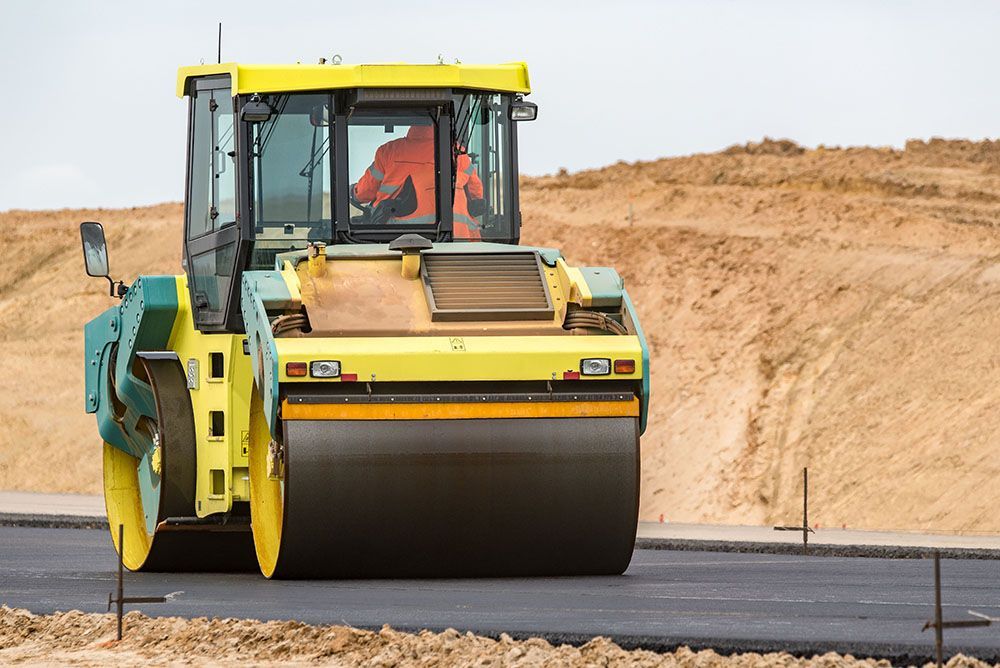 A yellow and green road roller is driving down a dirt road.