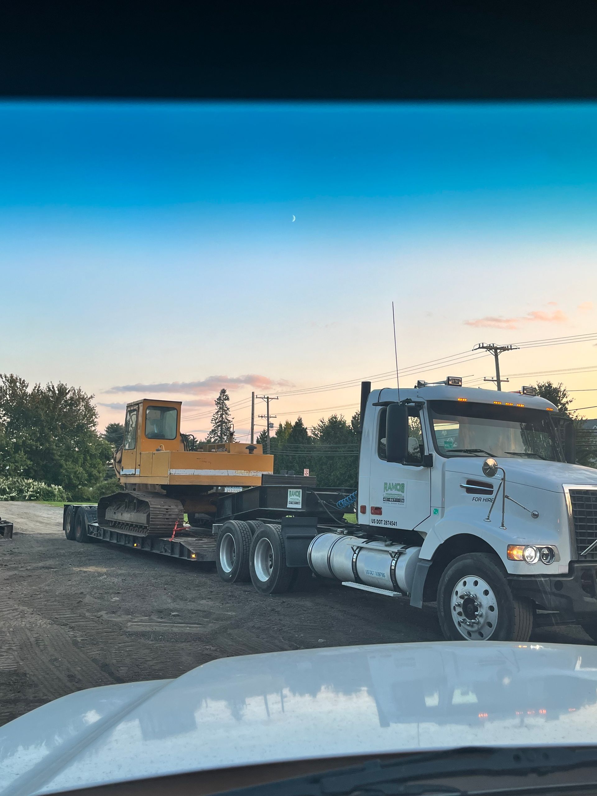 A white semi truck is carrying a yellow excavator on a trailer.
