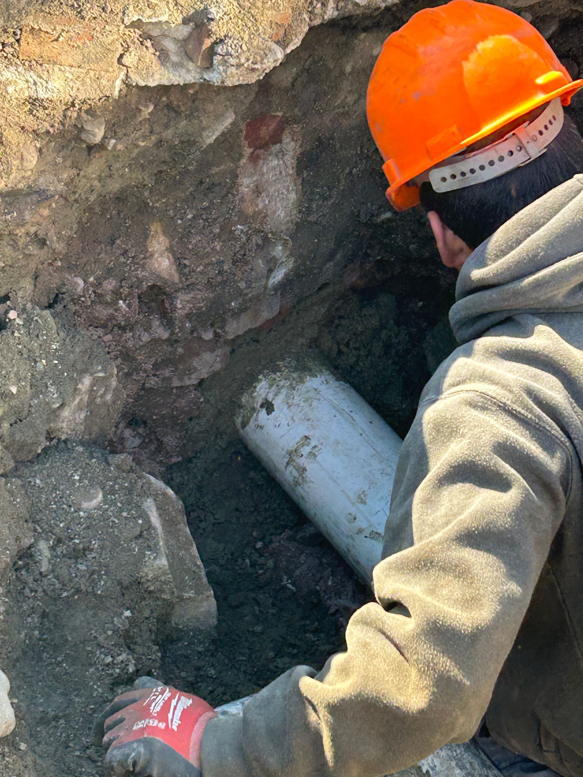 A man wearing an orange hard hat is working on a pipe in the dirt.