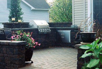 Outdoor kitchen with brick walls, grill, and cabinetry. Potted plants and brick patio.