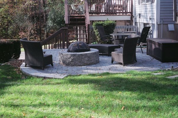 Circular patio with fire pit, chairs, and lawn, near a house and steps.