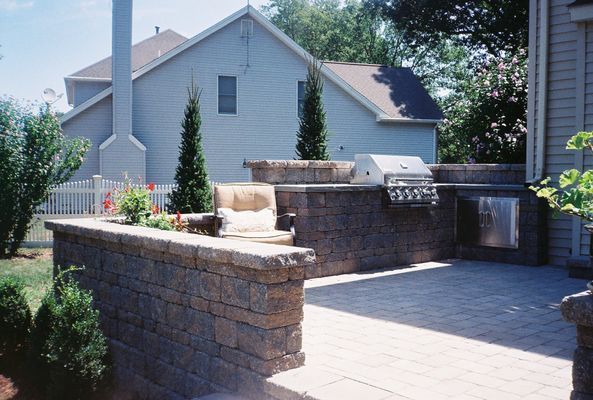 Stone patio with built-in grill and waterfall feature, beige brick walls, tall evergreens, and a beige chair.