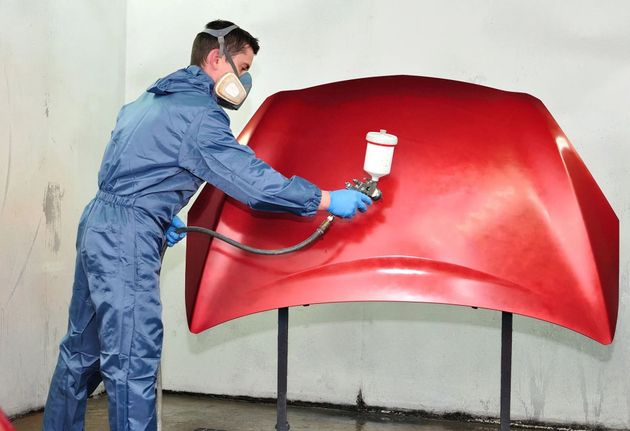 Person in protective gear spray-painting a large red car hood in a workshop