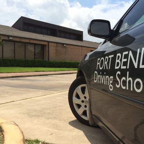 Black Fort Bend Driving School car parked outside a brick building on a sunny street.