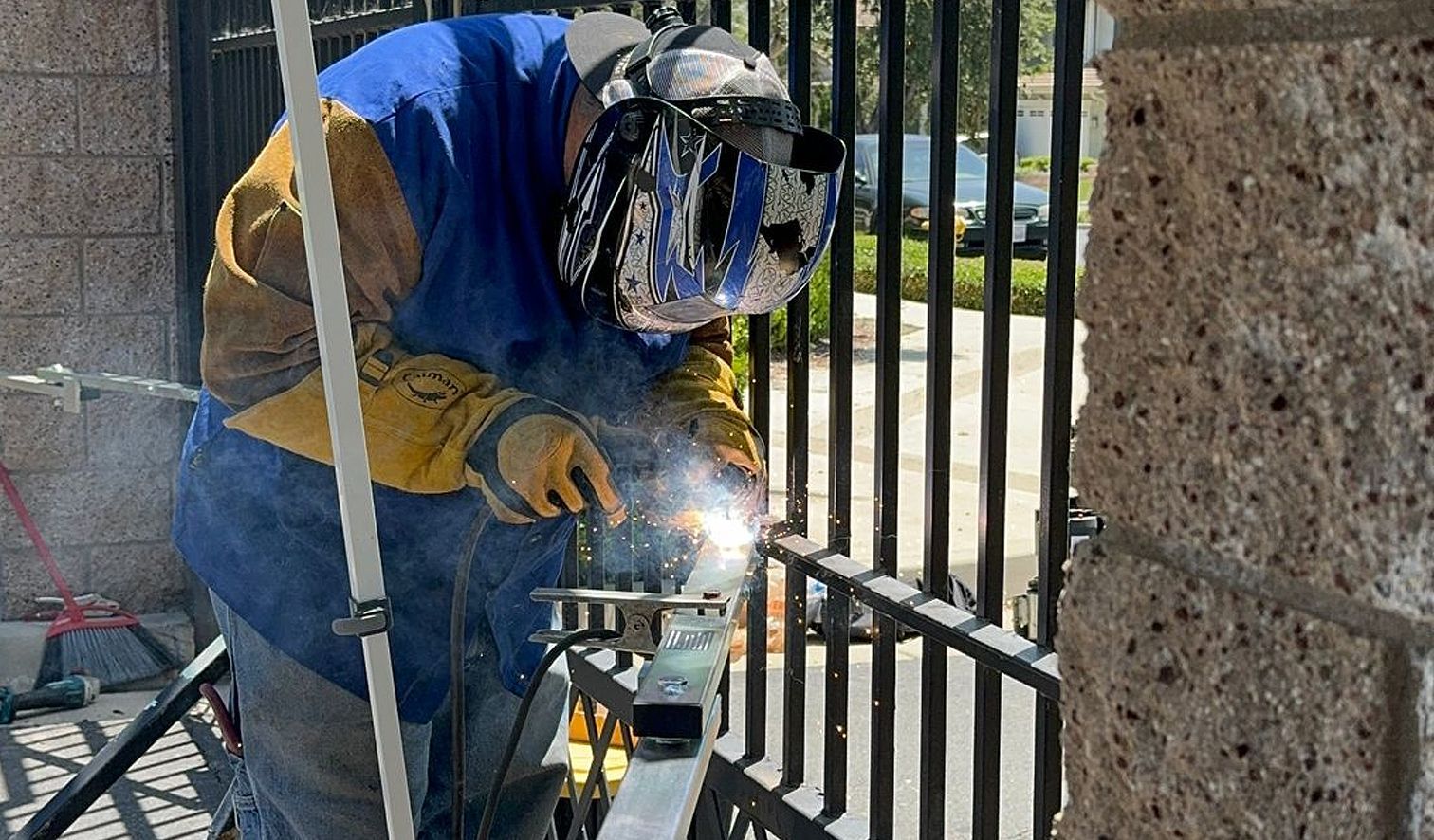 A man wearing a welding helmet is welding a metal fence.