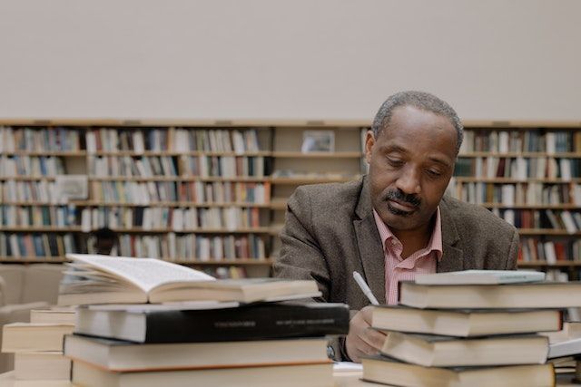 A black man works in a library, books piled around him on a table