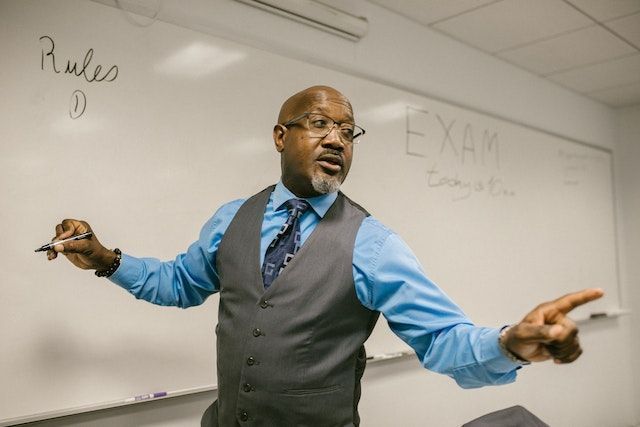 A black man instructs a class, pointing away from the white board towards them