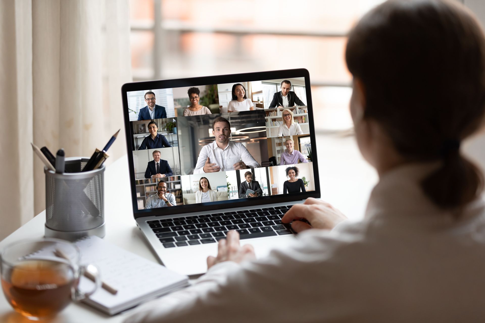 Image of a person watching an online webinar with several participants on a laptop computer. The person is sitting at a desk in front of a window.