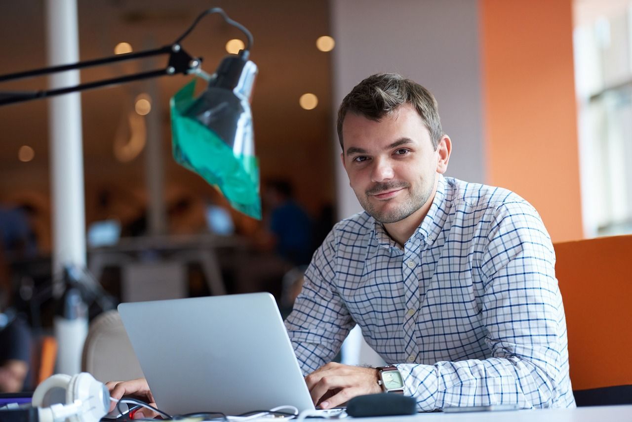 A man with short brown hair wearing a plaid shirt works on his laptop in an office