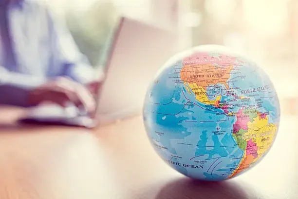 In the foreground, a globe sits on top of a table while a man works on a laptop in the background