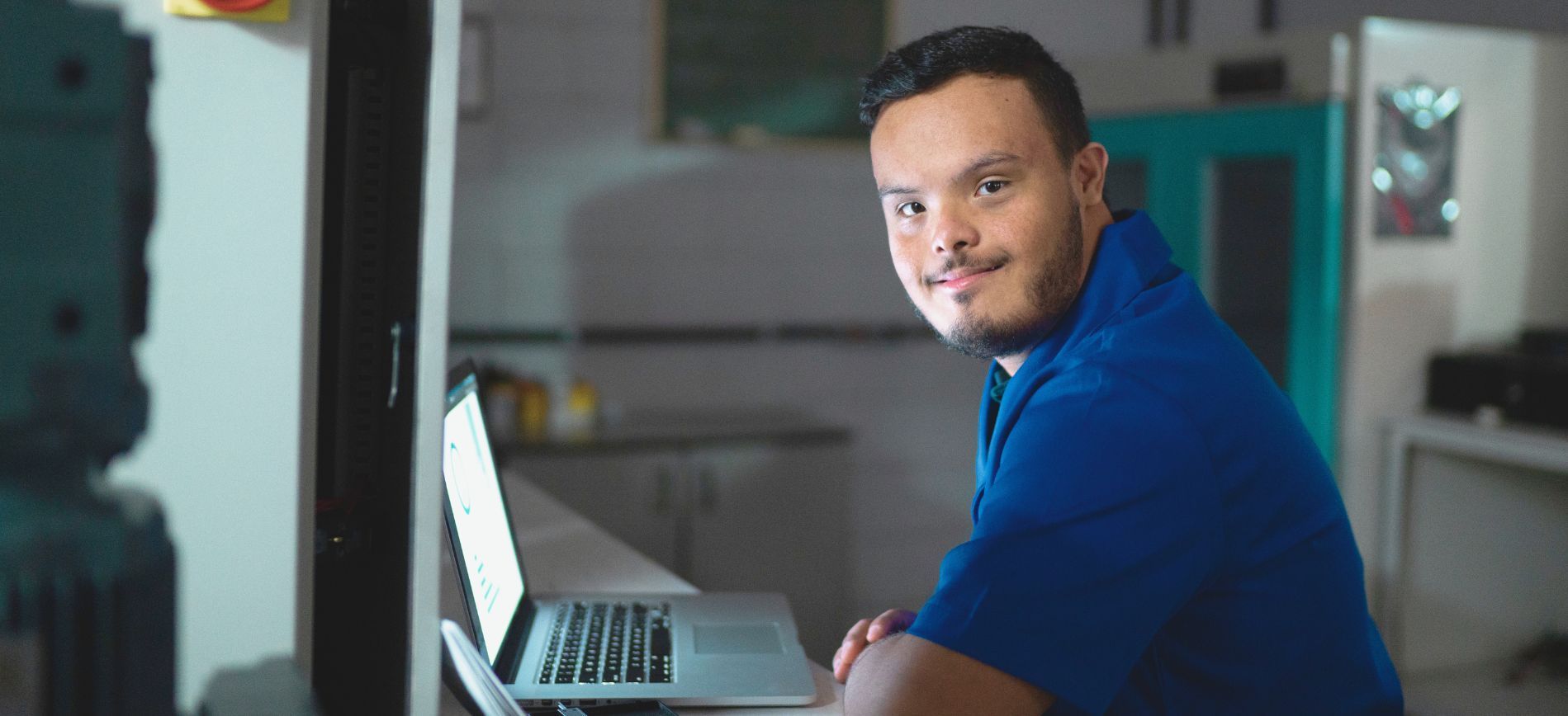 A white neurodiverse man wearing a blue shirt and working on a laptop smiles at the camera