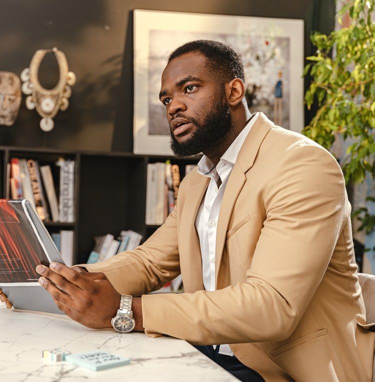A Black bearded man in a white button down and tan suit coat discusses a diversity magazine article with unseen persons in a conference room
