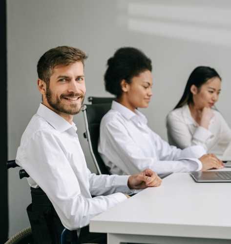 An white man using a wheelchair smiles at the camera, his laptop in front of him on the table